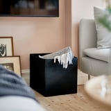 Schwarze DRÖNA Aufbewahrungsbox auf Holzboden, hellgraue Decke hängt heraus.