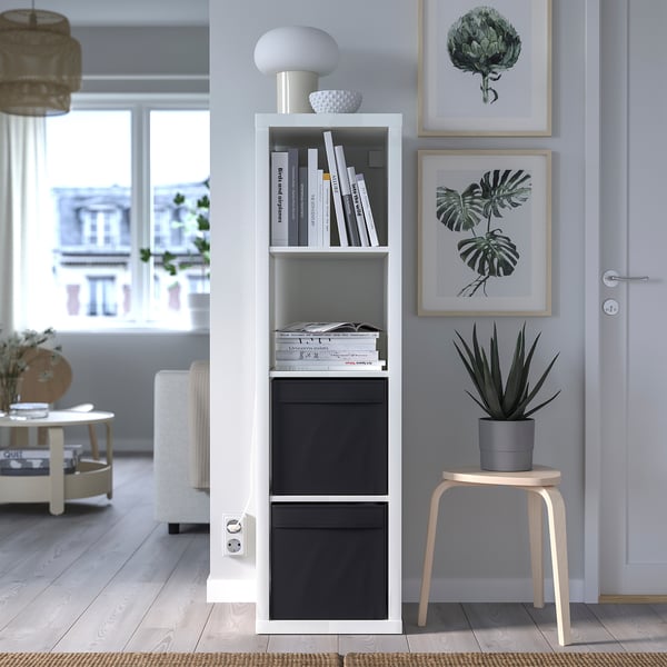 Modernes Wohnzimmer mit Kallax-Regalen, DRÖNA-Boxen, Lampe und Dekoration. Holzhocker mit Pflanze, Kunstdrucken und gemütlichem Teppich an großen Fenstern.