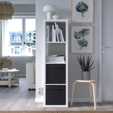 Modernes Wohnzimmer mit Kallax-Regalen, DRÖNA-Boxen, Lampe und Dekoration. Holzhocker mit Pflanze, Kunstdrucken und gemütlichem Teppich an großen Fenstern.