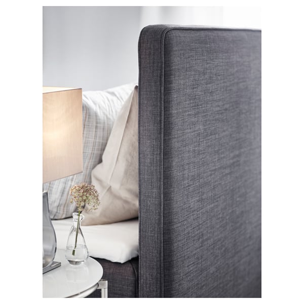 Modernes Bett mit weichem, grau gepolstertem Kopfteil. Weiße Lampe und kleine Vase mit Blumen auf dem Nachttisch, die eine gemütliche Schlafzimmerszene schaffen.
