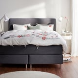 Modernes Bett mit grau gepolstertem Kopfteil, weißer Blumenbettdecke, zwei Lampen, gemütlicher Rahmen.