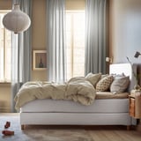 Schlafzimmer mit hellem DUNVIK-Bett, beigefarbener Bettwäsche, weißen Vorhängen und Hängelampe.