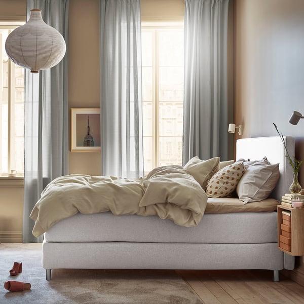 Ungemachtes Bett mit beiger Bettwäsche, Kopfteil aus Holz, stilvolle Hängelampe. Schlafzimmer großes Fenster mit Vorhängen und Blick auf die Gebäude.