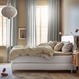 Ungemachtes Bett mit beiger Bettwäsche, Kopfteil aus Holz, stilvolle Hängelampe. Schlafzimmer großes Fenster mit Vorhängen und Blick auf die Gebäude.