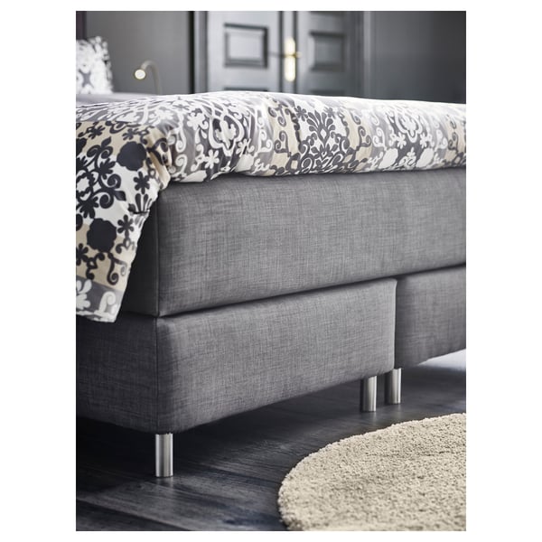 Modernes, graues Polsterbett mit gemusterter Decke, schlanke Metallbeine, auf Hartholzboden positioniert.