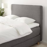 Ein grau gepolstertes Bett mit weichem Kopfteil mit sauberer weißer Bettwäsche und Kissen vor einer minimalistischen weißen Wand.