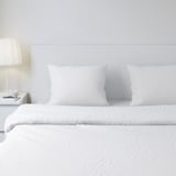 Weiße DVALA-Bettwäsche mit Baumwollkissenbezügen auf einem minimalistischen Bett, ruhige Atmosphäre, kleine Lampe.