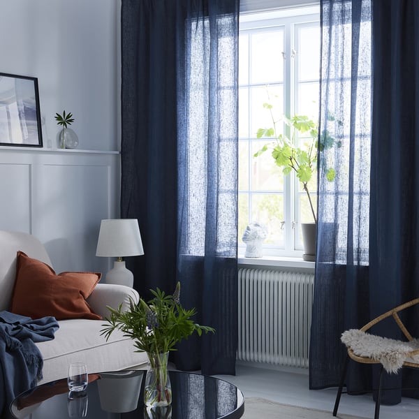 Gemütliches Wohnzimmer mit weißer Couch, rostigen Kissen, blauen Vorhängen, grüner Vase auf dem Tisch, geflochtenem Stuhl, weißem Teppich.