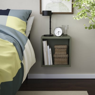 Kleines grünes EKET-Wandregal mit einer Lampe, einer Uhr und einem Korb neben einem Bett mit bunten Kissen. Sauberes, modernes Design.