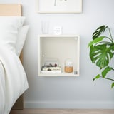 Minimalistisches Schlafzimmer mit weißem EKET-Würfelregal zur Aufbewahrung und Präsentation, das eine Uhr, Pflanzen und Accessoires zeigt und in der Nähe eines Bettes platziert ist.