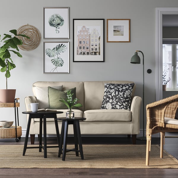 Modernes minimalistisches Wohnzimmer: beiges EKHOLMA-Sofa, schwarzer Beistelltisch, Korbstuhl, grauer Teppich, grüne Pflanzen, Wandkunst.