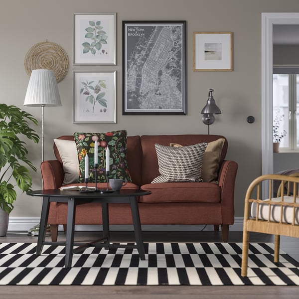 Modernes Wohnzimmer mit kleinem braunem EKHOLMA-Sofa, schwarzem Couchtisch, schwarz-weiß gestreiftem Teppich, bunten Kissen, Stehlampe, Wandkunst und Pflanze.