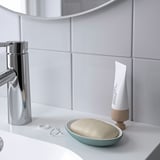 Eine minimalistische Badezimmertheke mit einer Seifenschale, einem Stück Seife in der Hand, neben einer Tube Sahne und einem Seifenspender.