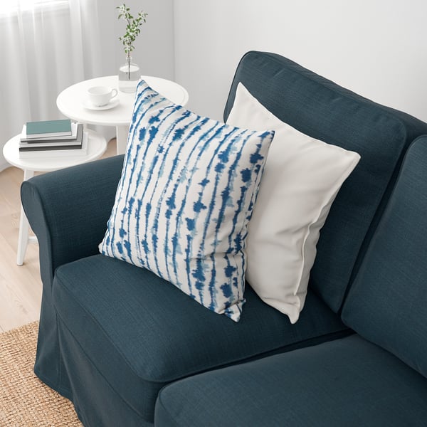 Ein gemütliches blaues Sofa mit gemusterten und weißen Kissen steht neben einem Tisch mit Büchern und Kaffeetassen.