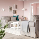 Ein gemütliches Wohnzimmer, ein grau-weiß gestreiftes EKTORP-Sofa mit einem kleinen Beistelltisch, einer Pflanze und Lampen in einem Raum mit rosa Wänden.
