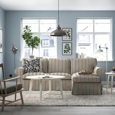 Gemütliches Wohnzimmer mit EKTORP-Sofa, Fenstern, Couchtisch, Akzentwand, Teppich, Drucken, Beistellstuhl, Lampe und Pflanze.