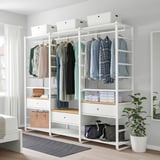 ELVARLI weißer Kleiderschrank mit Regalen und Schubladen bietet Platz für verschiedene Kleidungsstücke und Taschen, positioniert neben einem grauen Teppich und einem weißen Bett, mit einer Pflanze an der Seite.