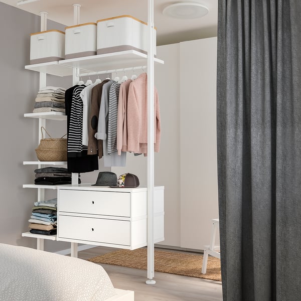 Weißer ELVARLI-Kleiderschrank mit Kleidung, Schubladen, Regalen und grauem Vorhang im aufgeräumten Schlafzimmer.