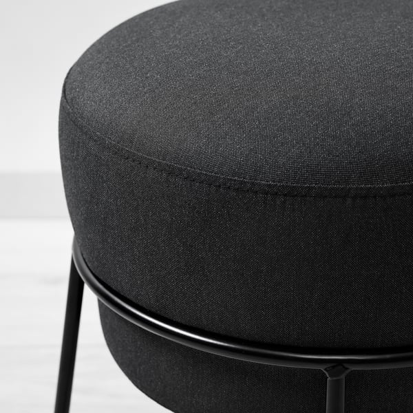 Runder gepolsterter Hocker mit dunkelgrauem Stoff über Metallgestell, der robust und modern wirkt.
