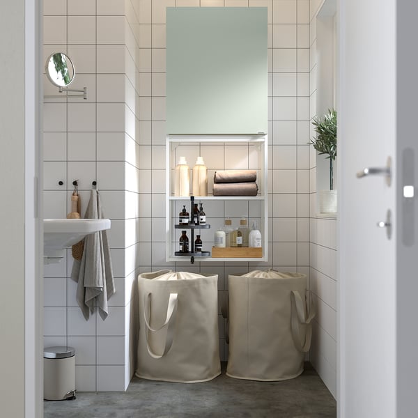 Modernes Badezimmer mit ENHET Stauraum. Weiße Fliesen, graugrüner Schrank, zwei Stoffkörbe. Pflanzen, Handtücher und Toilettenartikel ordentlich angeordnet.