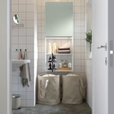 Modernes Badezimmer mit ENHET Stauraum. Weiße Fliesen, graugrüner Schrank, zwei Stoffkörbe. Pflanzen, Handtücher und Toilettenartikel ordentlich angeordnet.