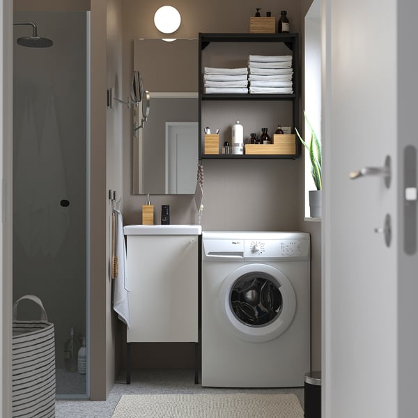 Eine moderne Waschküche und ein Badezimmer mit ENHET-Schrank, Frontlader-Waschmaschine, Waschbecken und offenen Regalen für Handtücher und Toilettenartikel. Minimalistisches Design.