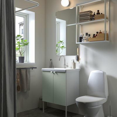 Modernes Badezimmer mit hellgrünem ENHET-Waschtisch, weißer Toilette, Spiegel mit Regalen. Handtücher und Pflanzen schmücken den ordentlich organisierten Raum.