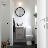 Kleines, modernes Badezimmer mit grauem Waschtisch ENHET, rundem Spiegel, weißer Toilette und Glasduschtür.