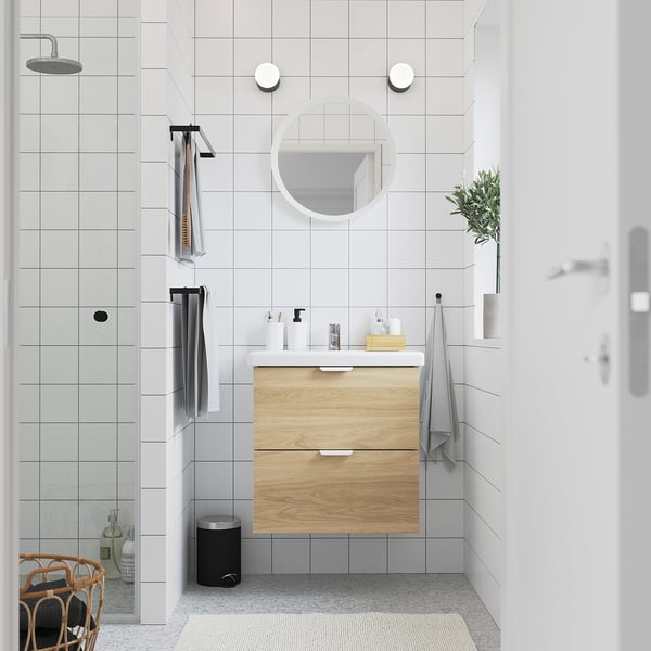 Modernes Badezimmer mit weißen Fliesen ENHET Waschtisch aus Holz. Schubladen aus hellem Holz, runder Spiegel, graue Handtücher und Pflanze. Kompakt und dennoch funktional.