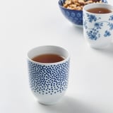 Zwei blau-weiß gemusterte Tassen mit Tee, eine geblümt, eine gepunktet, neben einer Schüssel mit Snacks.