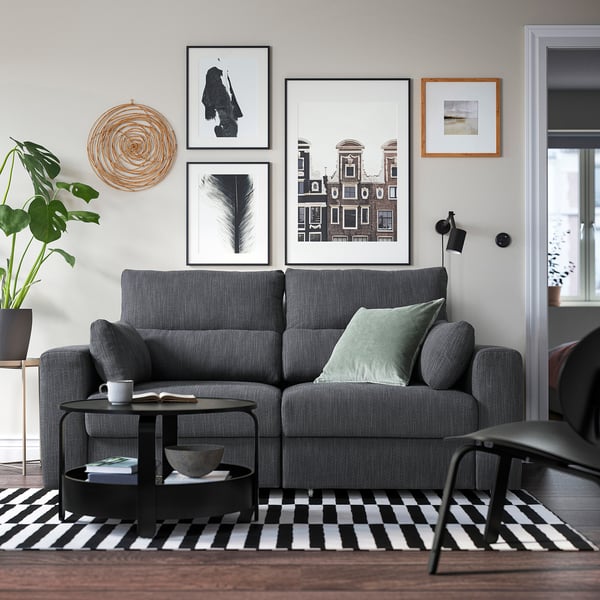 Modernes Wohnzimmer mit großem grauem ESKILSTUNA-Sofa, schwarzem Couchtisch, schwarz-weißem Teppich, Wandkunst und Pflanzen.