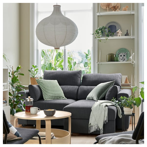 Modernes Wohnzimmer mit grauer Couchgarnitur, Couchtisch, gemütlichen Decken, grünen Kissen, Lampe, Pflanzen und Regalen.