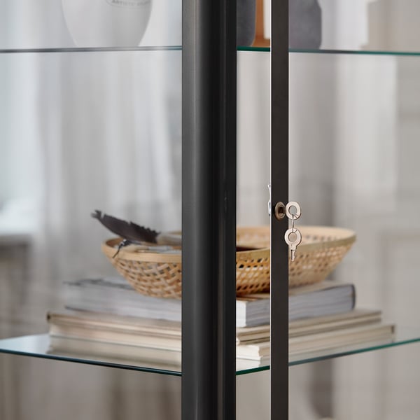 Moderner Schrank mit Glastür und schwarzem Rahmen, in dem Bücher und ein geflochtener Korb ausgestellt sind. Schlankes, robustes Design.