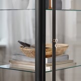 Moderner Schrank mit Glastür und schwarzem Rahmen, in dem Bücher und ein geflochtener Korb ausgestellt sind. Schlankes, robustes Design.