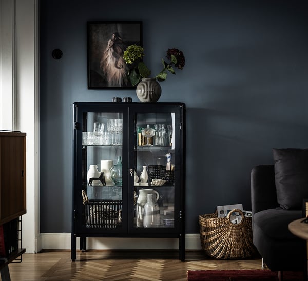 Schwarzer FABRIKÖR-Schrank mit Glastüren, in dem Vasen und Gläser ausgestellt sind, gekrönt von einer gestreiften Vase mit Blumen. Moderne, stilvolle Wohnzimmereinrichtung.