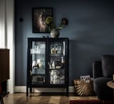 Schwarzer FABRIKÖR-Schrank mit Glastüren, in dem Vasen und Gläser ausgestellt sind, gekrönt von einer gestreiften Vase mit Blumen. Moderne, stilvolle Wohnzimmereinrichtung.