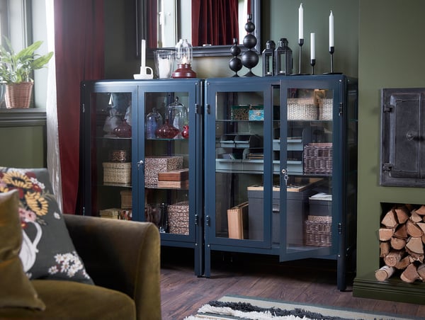 Wohnzimmer mit blauer FABRIKÖR Metall- und Glasvitrine mit Dekorationsgegenständen, daneben braune Couch mit Blumenkissen.