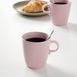 Zwei hellrosa Tassen, gefüllt mit Kaffee und Metalllöffeln, neben einem Teller mit einem Gebäck.
