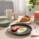 Teller mit Spiegelei und Tomaten, gestapelte Tassen, Croissants auf dem Tablett, Saft und Besteck auf dem Tisch.