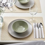 Gedeckter Tisch mit FÄRGKLAR Tellern, Schalen und Gläsern auf geflochtenen Matten, bereit zum Essen.