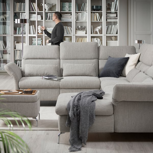 Modernes Wohnzimmer mit grauem FAMMARP-Sofa, verschiedenen Kissen und einem Mann, der mit Bücherregalen voller Bücher und Dekorationsgegenstände interagiert.