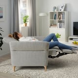 Frau, die sich auf einem Liegesessel entspannt, die Füße erhöht, in einem gemütlichen Wohnzimmer mit Büchern, Pflanzen und einem Fernseher.
