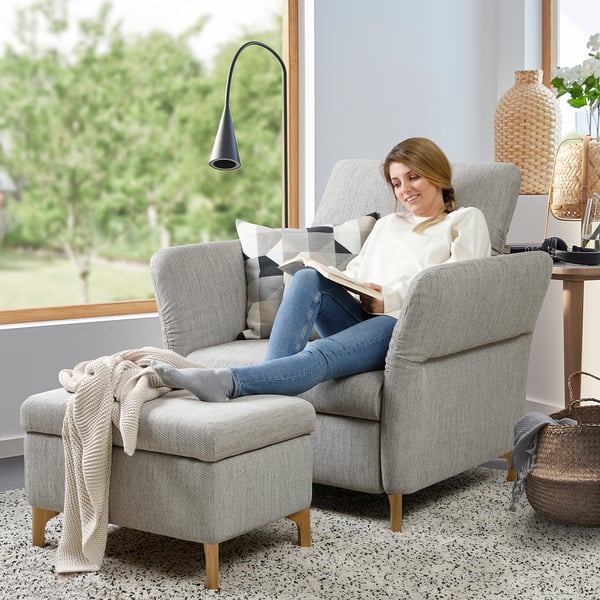 Frau liest in FAMMARP grauem Stuhl, weißem Pullover, blauen Jeans, gemütlichem Zimmer mit großem Fenster, Lampe und Korb.