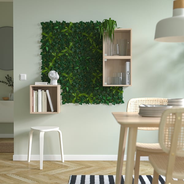 Modernes Wohnzimmer mit grüner Kunstpflanzenwand, schwebenden Regalen, weißem Hocker und Esstisch aus Holz mit Korbstühlen.