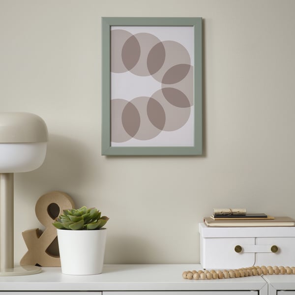 An einer Wand hängt ein grün gerahmtes Bild, umgeben von einem weißen Schreibtisch mit Dekorationsgegenständen wie einer Lampe, einer Pflanze und Holzperlen.