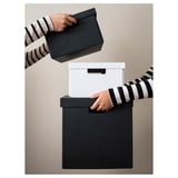 Eine Person mit schwarz-weißen Aufbewahrungsboxen. Die schwarze Box hat einen Griff, im Hintergrund ist eine weiße Box mit Deckel zu sehen.