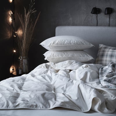 Weißes Bett mit FJÄLLBRÄCKA Bettdecke, zwei Kissen, kariertem Kissenbezug, Trockenblumen, Wandleuchten.