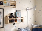 Kinderzimmer mit weißen FLISAT-Holzregalen mit Büchern und Puppen, blauen Akzenten, einer Wandleuchte und einem Bett mit blau-weiß gestreiften Kissen.