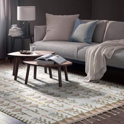 Wohnzimmerszene: graues Sofa mit pastellfarbenen Kissen, zwei hölzerne Couchtische, bunter Teppich, Lampe mit Büchern und Decke. Teppiche bedecken hauptsächlich den Boden.