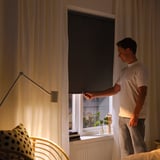 Mann stellt schwarzes Rollo im Schlafzimmer ein und enthüllt Fenster mit Pflanzen und Büchern.